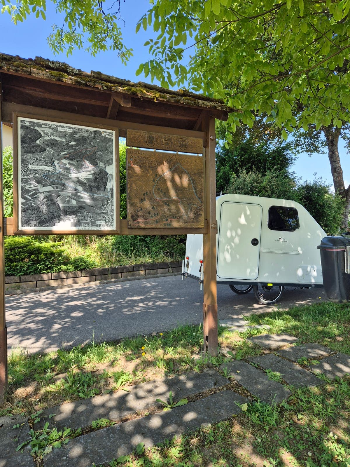 Der weiße Tiny-Camper steht am Wegesrand neben einer hölzernen Wanderkarte im Grünen
